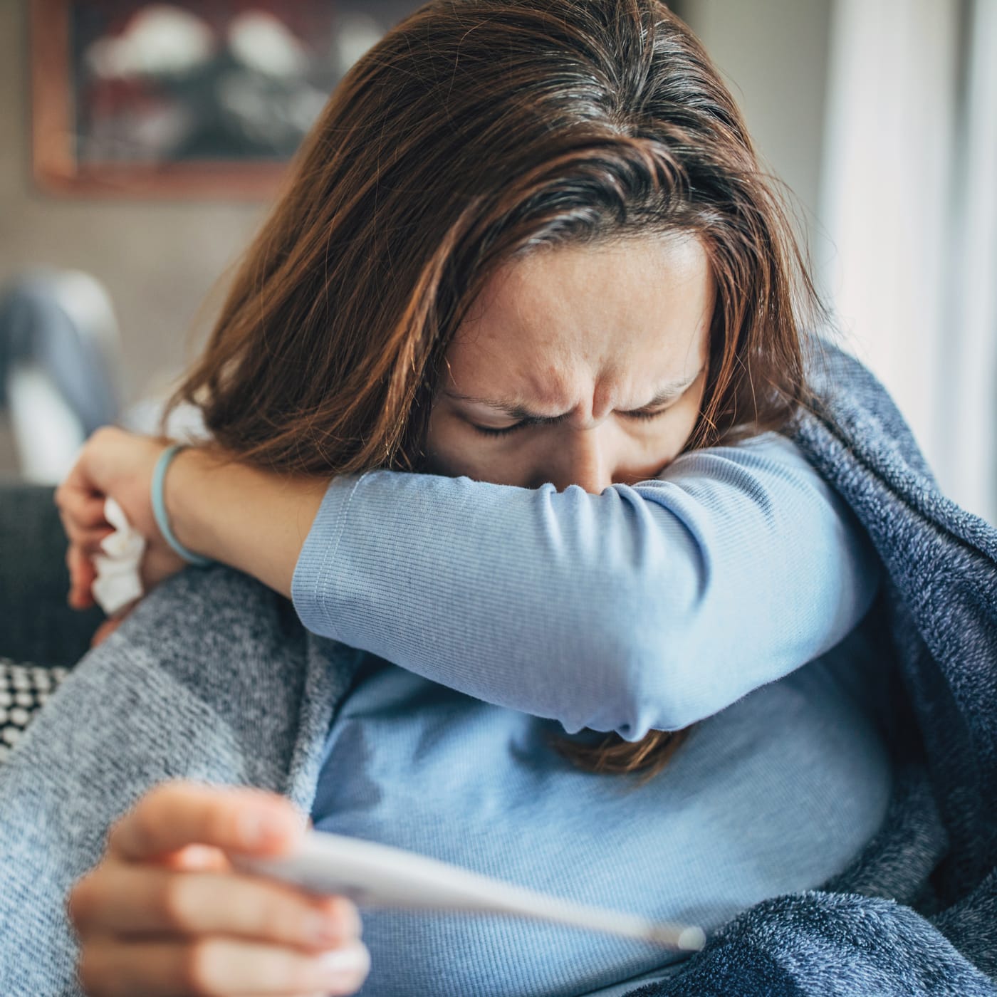 A person holding a thermometer coughing into their arm